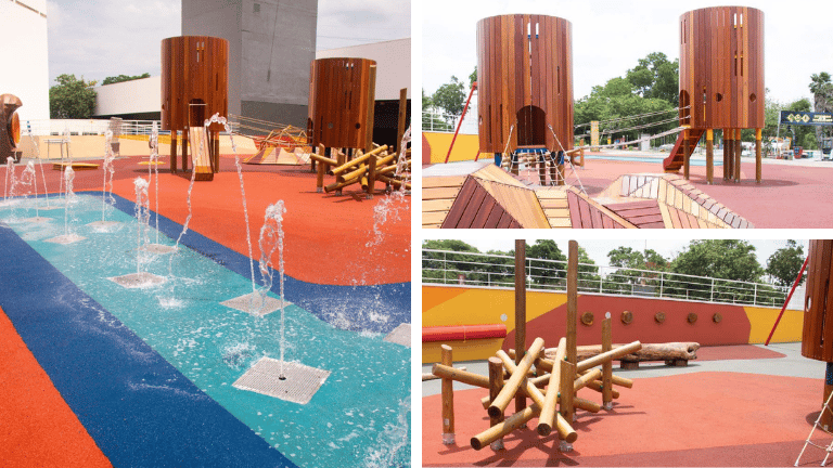 Playground ao ar livre com piso emborrachado colorido, torres de madeira, circuito de equilíbrio e área com fontes de água.